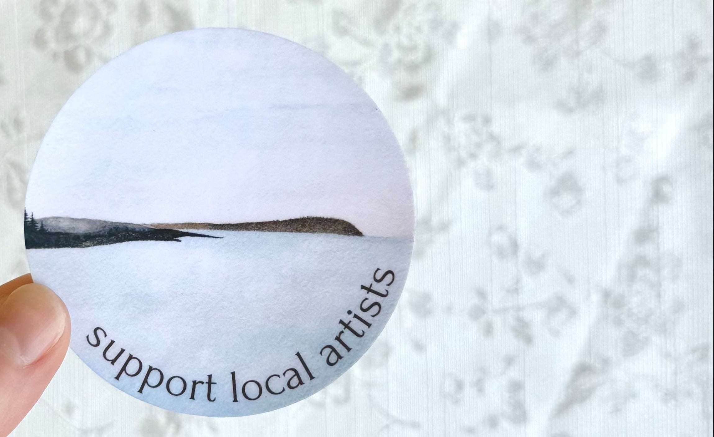 Hand holding a round sticker with 'support local artists' text on a textured white background